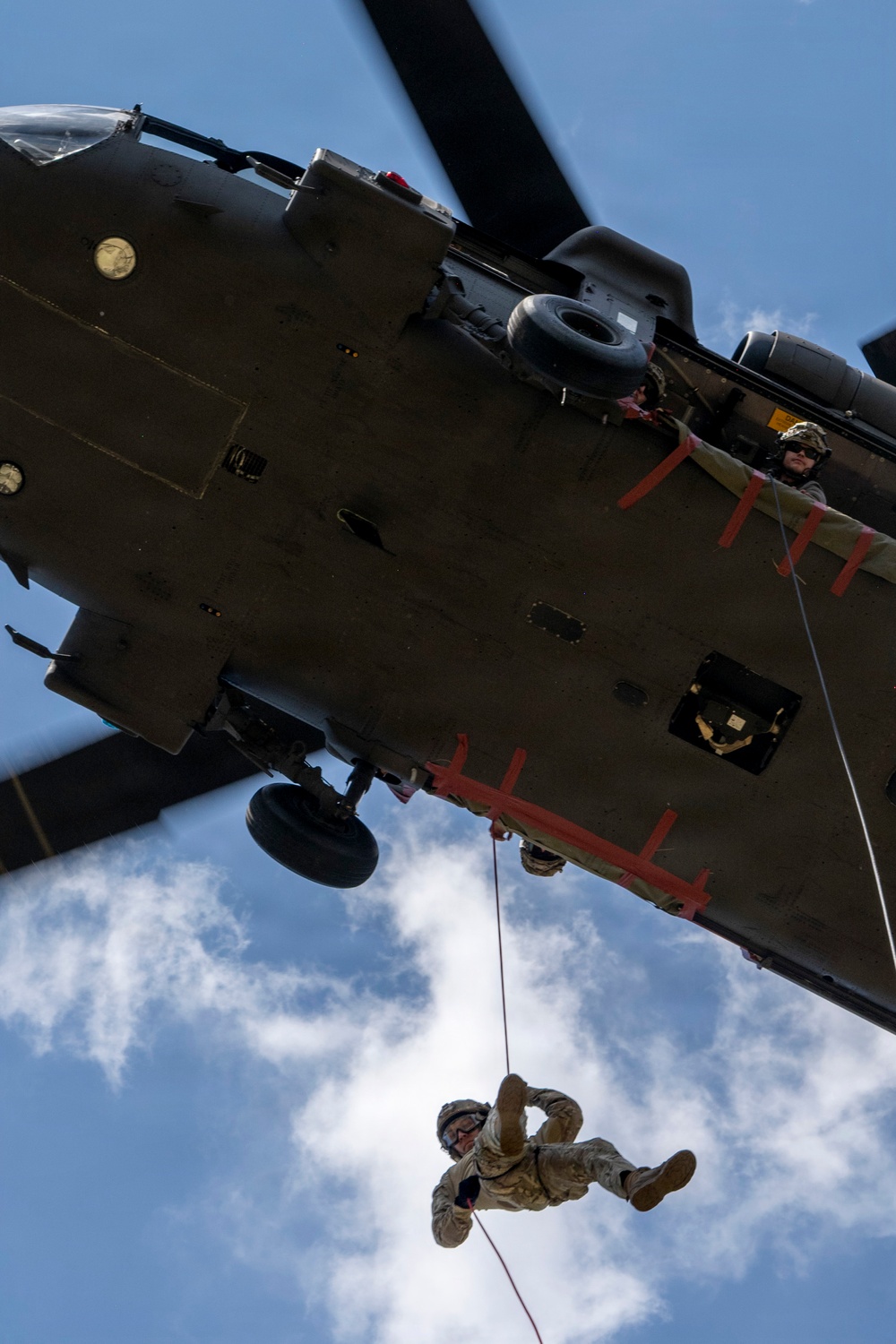 A military helicopter, viewed from below, with a soldier rappelling down a rope from the aircraft. A crew member is visible leaning out of the open door observing the descent. The image is taken from a ground-up perspective against a partly cloudy blue sky.