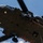TACP Airmen master vertical infiltration A military helicopter, viewed from below, with a soldier rappelling down a rope from the aircraft. A crew member is visible leaning out of the open door observing the descent. The image is taken from a ground-up perspective against a partly cloudy blue sky.