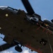 A military helicopter, viewed from below, with a soldier rappelling down a rope from the aircraft. A crew member is visible leaning out of the open door observing the descent. The image is taken from a ground-up perspective against a partly cloudy blue sky.