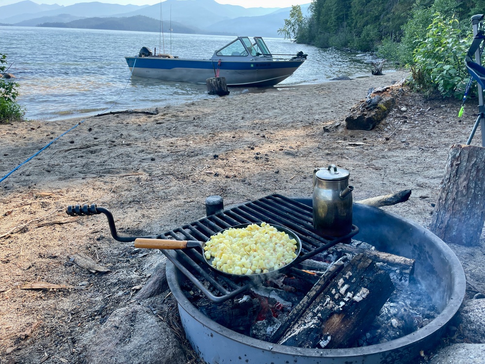 A campfire cooking scene on a sandy lakeshore beach, with a frying pan containing diced potatoes or similar food and a metal kettle/pot heating on a cast iron grill grate over an open fire. In the background, a blue and silver motorboat is beached on the shoreline of a calm lake surrounded by forested mountains. The scene depicts a wilderness camping or boating trip with outdoor cooking.