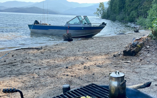 A campfire cooking scene on a sandy lakeshore beach, with a frying pan containing diced potatoes or similar food and a metal kettle/pot heating on a cast iron grill grate over an open fire. In the background, a blue and silver motorboat is beached on the shoreline of a calm lake surrounded by forested mountains. The scene depicts a wilderness camping or boating trip with outdoor cooking.