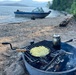 A campfire cooking scene on a sandy lakeshore beach, with a frying pan containing diced potatoes or similar food and a metal kettle/pot heating on a cast iron grill grate over an open fire. In the background, a blue and silver motorboat is beached on the shoreline of a calm lake surrounded by forested mountains. The scene depicts a wilderness camping or boating trip with outdoor cooking.