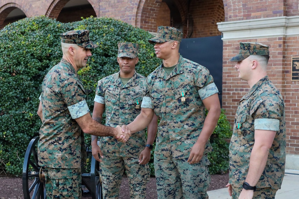 Four U.S. Marines in MARPAT digital camouflage uniforms are gathered outside a brick building. A senior Marine on the left is shaking hands with a taller Marine identified by a name tape reading 'WILSON,' while two other Marines stand nearby. Three of the four Marines are wearing green medals/ribbons on their uniforms, suggesting a recent award or recognition ceremony.