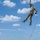 TACP Airmen master vertical infiltration A military or tactical training exercise is underway in an open field, where one person in camouflage uniform is being lifted high into the air via a red rope attached to a harness, likely connected to a helicopter above the frame. A group of approximately seven personnel on the ground are managing the rope and observing the extraction. The setting appears to be a rural training area with green hills and a partly cloudy sky.