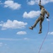 A military or tactical training exercise is underway in an open field, where one person in camouflage uniform is being lifted high into the air via a red rope attached to a harness, likely connected to a helicopter above the frame. A group of approximately seven personnel on the ground are managing the rope and observing the extraction. The setting appears to be a rural training area with green hills and a partly cloudy sky.