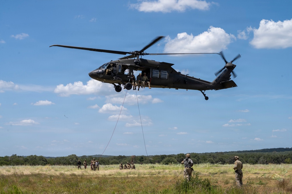 A military UH-60 Black Hawk helicopter hovers low over an open grassy field during what appears to be a rappelling or fast-rope training exercise. Several soldiers are visible seated in the open door of the helicopter with a rope descending to the ground, while additional personnel on the ground observe and manage the operation. The setting appears to be a rural training area with tree lines in the background under a partly cloudy sky.