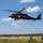 TACP Airmen master vertical infiltration A military UH-60 Black Hawk helicopter hovers low over an open grassy field during what appears to be a rappelling or fast-rope training exercise. Several soldiers are visible seated in the open door of the helicopter with a rope descending to the ground, while additional personnel on the ground observe and manage the operation. The setting appears to be a rural training area with tree lines in the background under a partly cloudy sky.