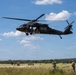 A military UH-60 Black Hawk helicopter hovers low over an open grassy field during what appears to be a rappelling or fast-rope training exercise. Several soldiers are visible seated in the open door of the helicopter with a rope descending to the ground, while additional personnel on the ground observe and manage the operation. The setting appears to be a rural training area with tree lines in the background under a partly cloudy sky.
