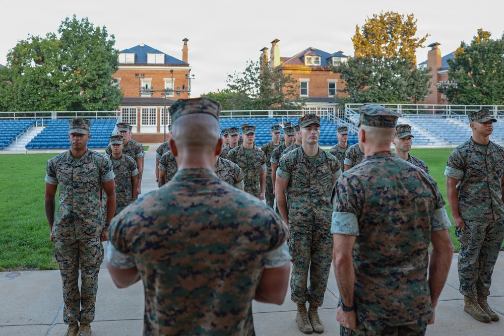 A formation of U.S. Marines in woodland MARPAT camouflage uniforms stands at attention on a parade field or athletic ground. Two Marines with their backs to the camera appear to be addressing or inspecting the assembled group. The setting includes brick colonial-style buildings and blue stadium bleachers in the background, suggesting a military installation or academy grounds.