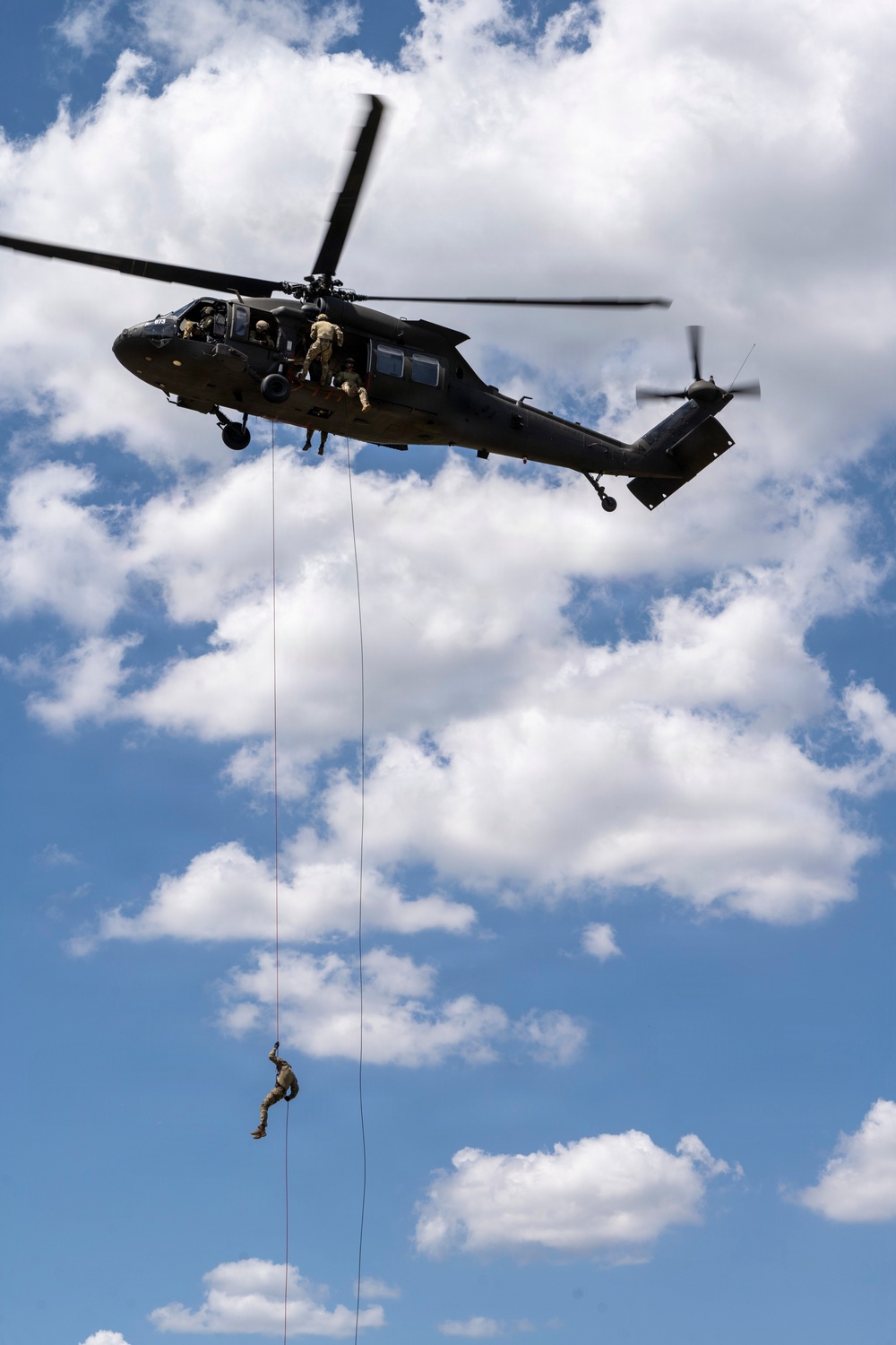 A military helicopter, consistent with a UH-60 Black Hawk, is hovering in a partly cloudy blue sky while personnel conduct a fast-rope or rappelling operation. At least one soldier is suspended mid-air on a rope well below the aircraft, while additional personnel are visible at the helicopter's door. This appears to be a military training or insertion exercise.