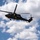 TACP Airmen master vertical infiltration A military helicopter, consistent with a UH-60 Black Hawk, is hovering in a partly cloudy blue sky while personnel conduct a fast-rope or rappelling operation. At least one soldier is suspended mid-air on a rope well below the aircraft, while additional personnel are visible at the helicopter's door. This appears to be a military training or insertion exercise.