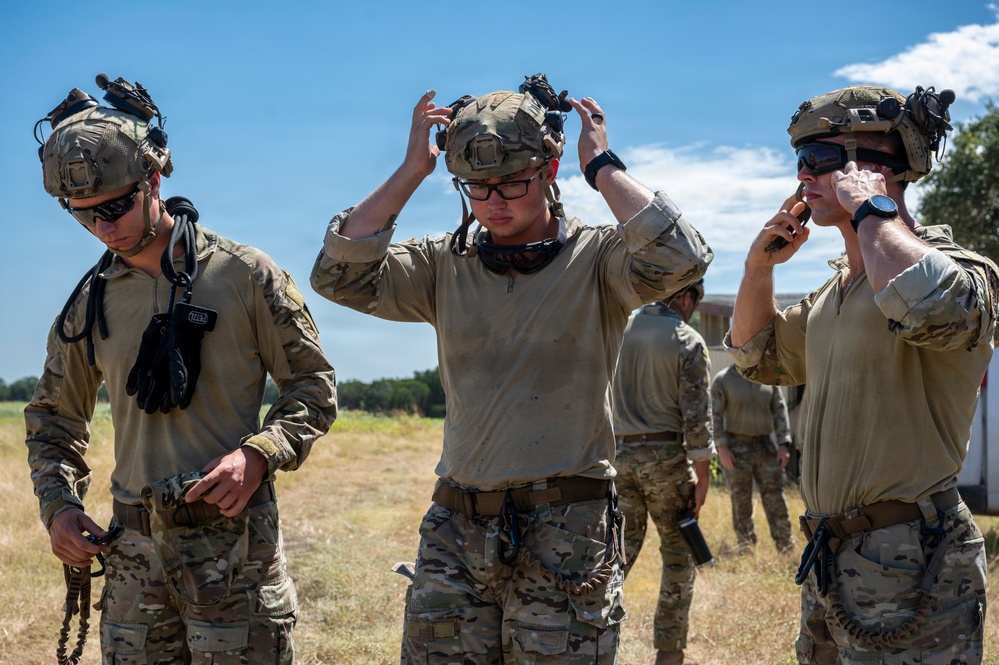 A group of military personnel in MultiCam/OCP camouflage uniforms are gathered in an open field under a clear blue sky. Three soldiers are prominently visible in the foreground, adjusting their tactical helmets and gear after what appears to be a training exercise. Their uniforms show signs of dirt and wear, suggesting recent physical activity.