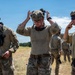 A group of military personnel in MultiCam/OCP camouflage uniforms are gathered in an open field under a clear blue sky. Three soldiers are prominently visible in the foreground, adjusting their tactical helmets and gear after what appears to be a training exercise. Their uniforms show signs of dirt and wear, suggesting recent physical activity.