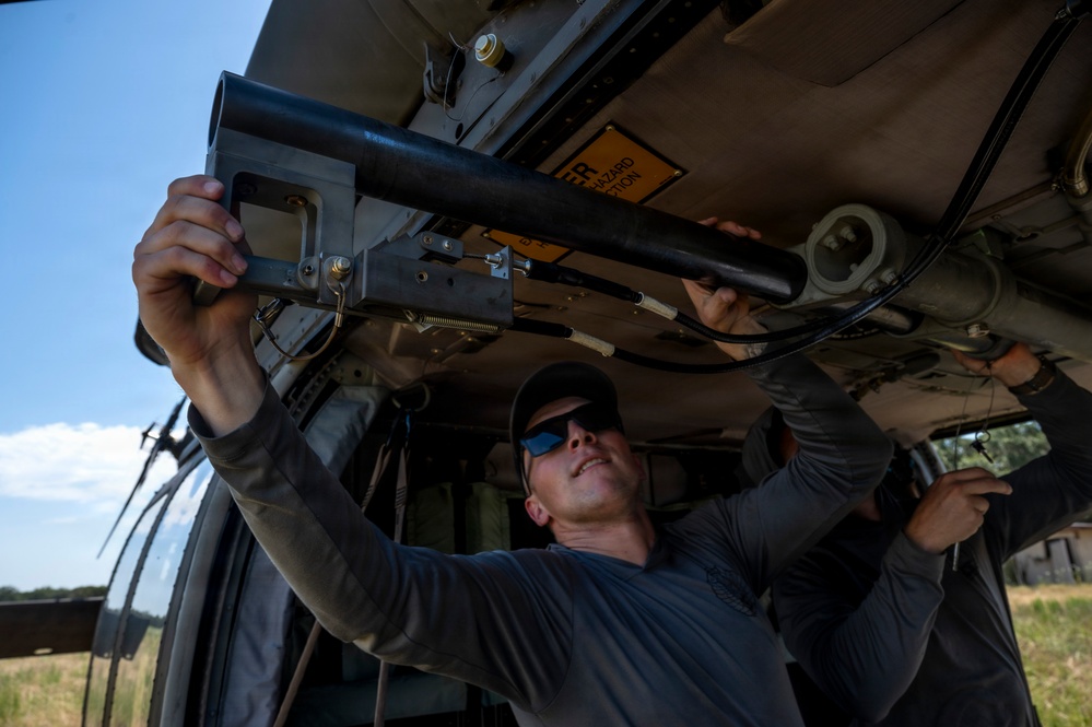 A person wearing sunglasses and a dark cap is working underneath what appears to be a military or utility helicopter, installing or adjusting a weapon mount or equipment bracket. The work is being performed outdoors in a field setting under clear blue skies. A second individual is partially visible in the background assisting with the task.