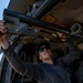 A person wearing sunglasses and a dark cap is working underneath what appears to be a military or utility helicopter, installing or adjusting a weapon mount or equipment bracket. The work is being performed outdoors in a field setting under clear blue skies. A second individual is partially visible in the background assisting with the task.