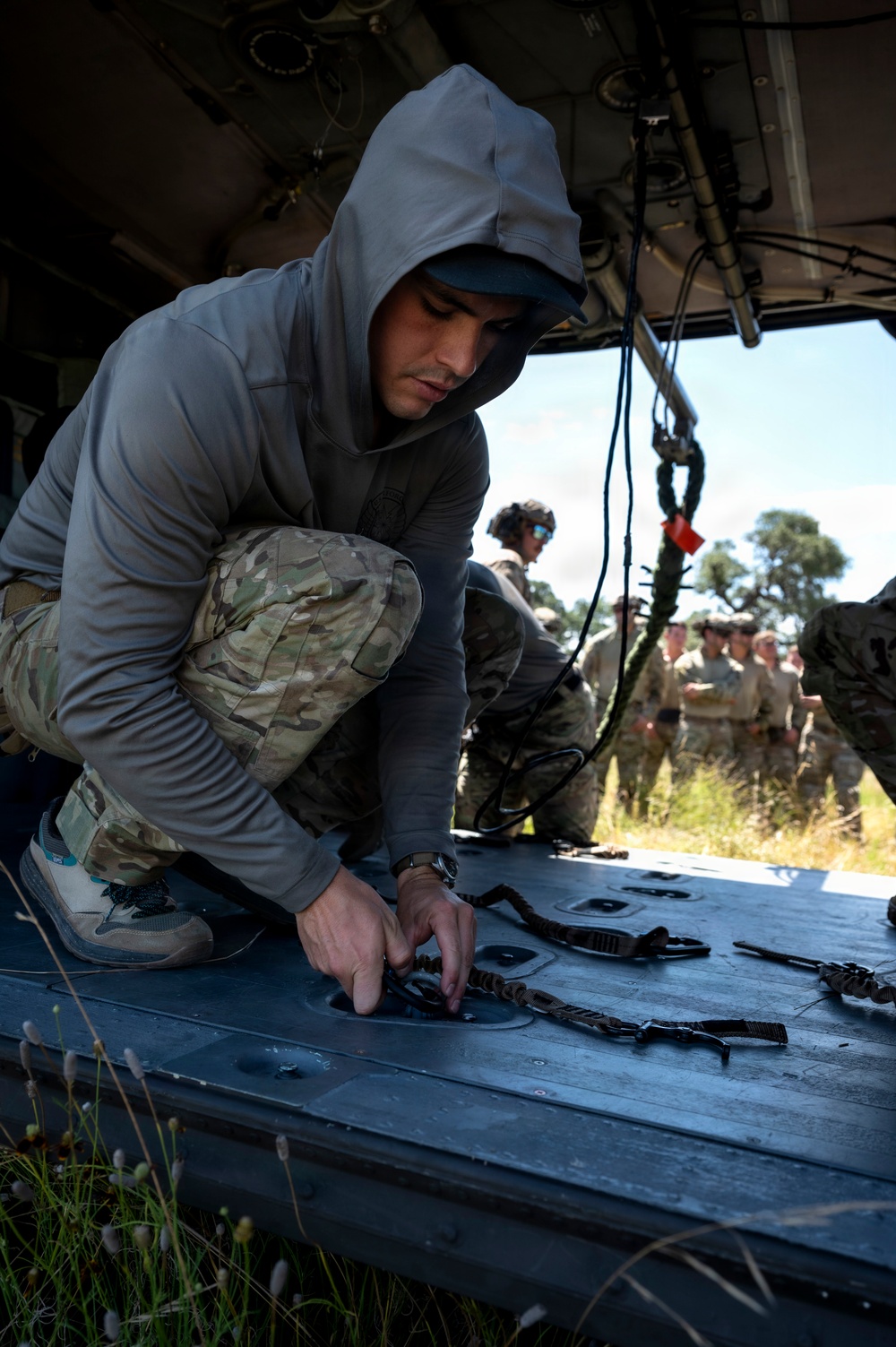 A man wearing a gray hoodie and multicam/OCP camouflage pants is kneeling inside what appears to be a military helicopter (likely a CH-47 Chinook or similar), handling straps or rigging equipment on the floor ramp. In the background, a group of military personnel in uniform are observing, suggesting this is a training or demonstration scenario. The setting appears to be an outdoor field environment.