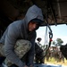 A man wearing a gray hoodie and multicam/OCP camouflage pants is kneeling inside what appears to be a military helicopter (likely a CH-47 Chinook or similar), handling straps or rigging equipment on the floor ramp. In the background, a group of military personnel in uniform are observing, suggesting this is a training or demonstration scenario. The setting appears to be an outdoor field environment.