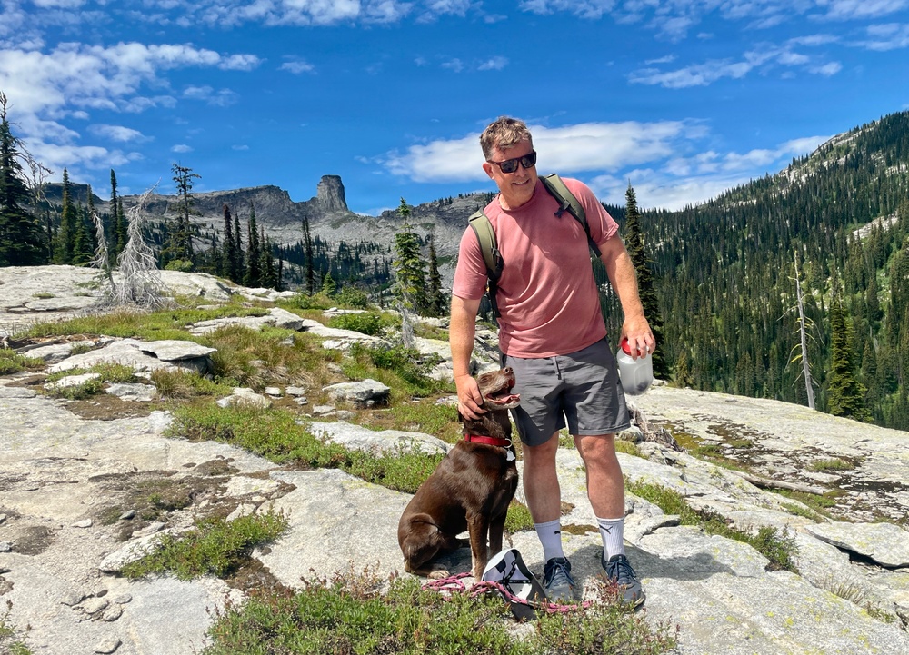 A man wearing a pink t-shirt, gray shorts, and sunglasses stands on a granite rock outcropping with a chocolate Labrador Retriever wearing a red collar. He is holding a water bottle and appears to be on a mountain hiking trail. The background features coniferous forest, dramatic rocky peaks, and a blue sky with scattered clouds.