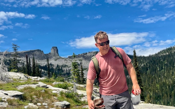 A man wearing a pink t-shirt, gray shorts, and sunglasses stands on a granite rock outcropping with a chocolate Labrador Retriever wearing a red collar. He is holding a water bottle and appears to be on a mountain hiking trail. The background features coniferous forest, dramatic rocky peaks, and a blue sky with scattered clouds.