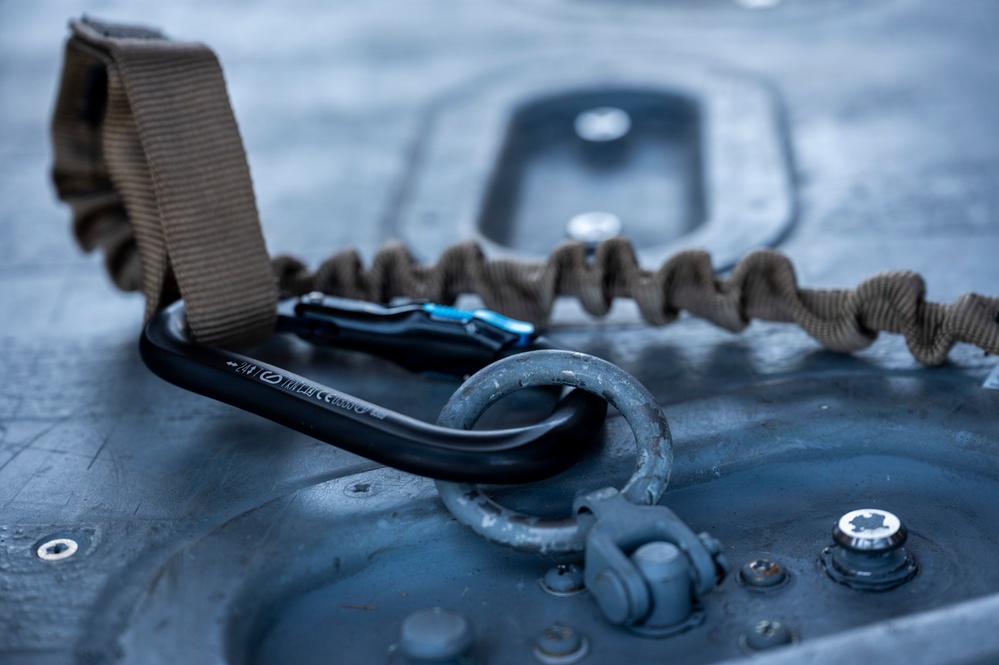 A close-up photograph of a black carabiner with a blue locking mechanism attached to a metal D-ring anchor point on a gray metallic surface. A tan/coyote brown bungee retention lanyard or safety tether connects to the carabiner. The image appears to be taken in a military or tactical equipment context, possibly on an aircraft or vehicle surface.