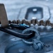 A close-up photograph of a black carabiner with a blue locking mechanism attached to a metal D-ring anchor point on a gray metallic surface. A tan/coyote brown bungee retention lanyard or safety tether connects to the carabiner. The image appears to be taken in a military or tactical equipment context, possibly on an aircraft or vehicle surface.