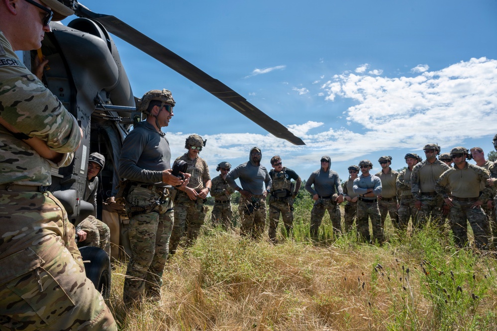 A group of military personnel are gathered in an open field next to a helicopter, appearing to receive a briefing or instruction from an individual standing near the aircraft. The personnel are wearing a mix of camouflage uniforms and gray athletic shirts, with tactical gear and helmets. The setting appears to be an outdoor training exercise or operational preparation.