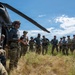 A group of military personnel are gathered in an open field next to a helicopter, appearing to receive a briefing or instruction from an individual standing near the aircraft. The personnel are wearing a mix of camouflage uniforms and gray athletic shirts, with tactical gear and helmets. The setting appears to be an outdoor training exercise or operational preparation.