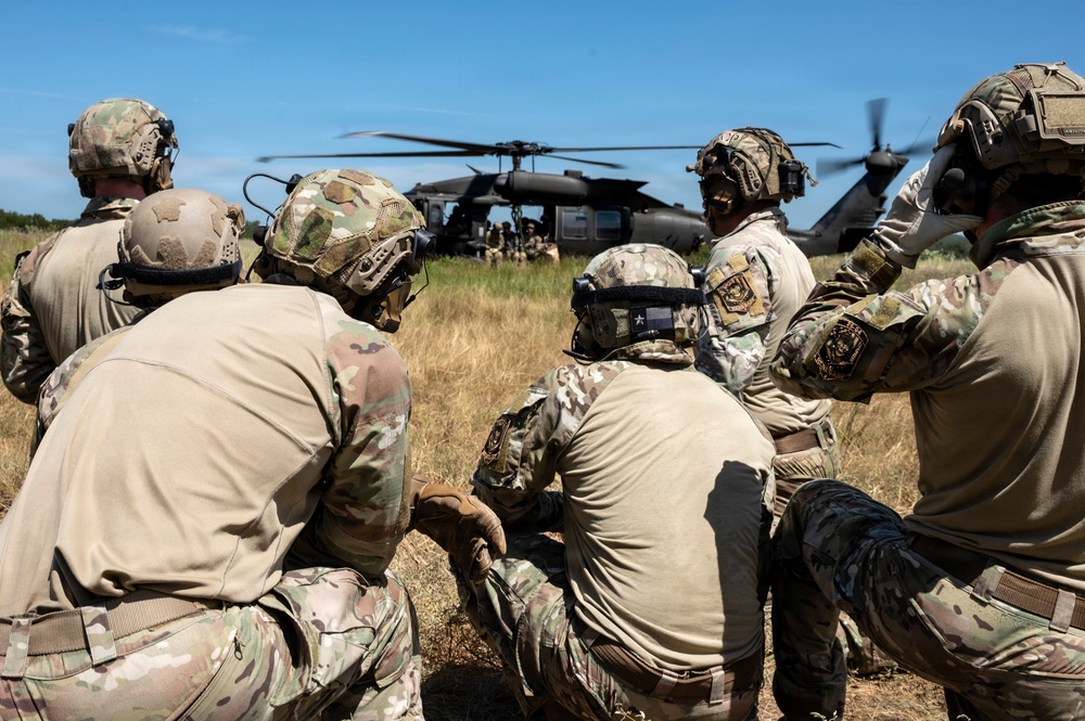 A group of military personnel in full combat gear are crouched in a grassy field observing a helicopter, likely a UH-60 Black Hawk, that has landed or is hovering nearby. The soldiers are wearing multicam/OCP camouflage uniforms, tactical helmets with communications equipment, and body armor. The scene appears to depict a training exercise or operational insertion/extraction scenario.