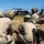TACP Airmen master vertical infiltration A group of military personnel in full combat gear are crouched in a grassy field observing a helicopter, likely a UH-60 Black Hawk, that has landed or is hovering nearby. The soldiers are wearing multicam/OCP camouflage uniforms, tactical helmets with communications equipment, and body armor. The scene appears to depict a training exercise or operational insertion/extraction scenario.