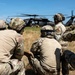 A group of military personnel in full combat gear are crouched in a grassy field observing a helicopter, likely a UH-60 Black Hawk, that has landed or is hovering nearby. The soldiers are wearing multicam/OCP camouflage uniforms, tactical helmets with communications equipment, and body armor. The scene appears to depict a training exercise or operational insertion/extraction scenario.