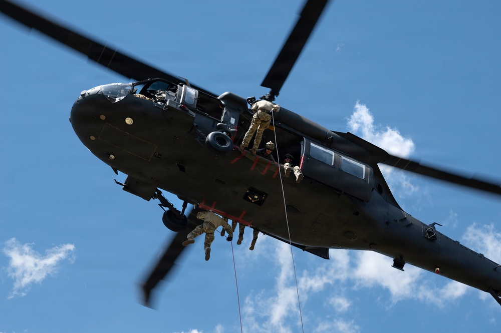 A military helicopter, consistent with a UH-60 Black Hawk, is photographed from below against a blue sky with scattered clouds. Multiple personnel in military/tactical uniforms are visible rappelling or fast-roping from the aircraft's side door. The image captures an active aerial insertion or training exercise in progress.