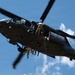 A military helicopter, consistent with a UH-60 Black Hawk, is photographed from below against a blue sky with scattered clouds. Multiple personnel in military/tactical uniforms are visible rappelling or fast-roping from the aircraft's side door. The image captures an active aerial insertion or training exercise in progress.