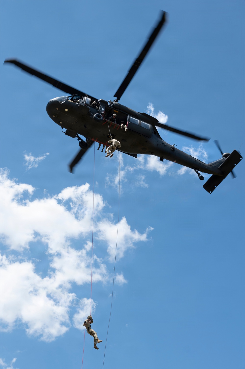 A military helicopter, identifiable as a UH-60 Black Hawk, is captured in flight against a partly cloudy blue sky. Two personnel are visible rappelling or fast-roping down from the aircraft on separate ropes, with one figure near the helicopter door and another significantly lower on the rope. This appears to be a military training or demonstration exercise involving helicopter insertion/extraction techniques.