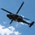 TACP Airmen master vertical infiltration A military helicopter, identifiable as a UH-60 Black Hawk, is captured in flight against a partly cloudy blue sky. Two personnel are visible rappelling or fast-roping down from the aircraft on separate ropes, with one figure near the helicopter door and another significantly lower on the rope. This appears to be a military training or demonstration exercise involving helicopter insertion/extraction techniques.