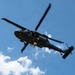 A military helicopter, identifiable as a UH-60 Black Hawk, is captured in flight against a partly cloudy blue sky. Two personnel are visible rappelling or fast-roping down from the aircraft on separate ropes, with one figure near the helicopter door and another significantly lower on the rope. This appears to be a military training or demonstration exercise involving helicopter insertion/extraction techniques.
