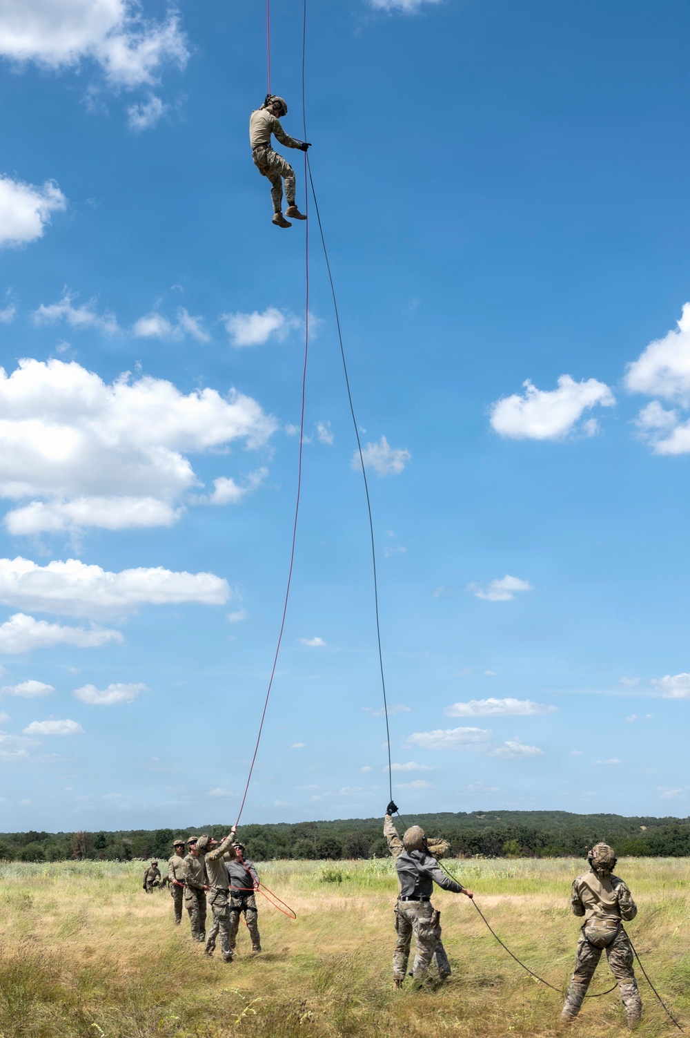 A military training exercise is underway in an open grassy field, with one soldier suspended high in the air on a rope, likely rappelling or being hoisted from a helicopter above the frame. Several personnel on the ground are managing guide ropes and safety lines. The setting appears to be a daytime outdoor training area with treeline in the background.