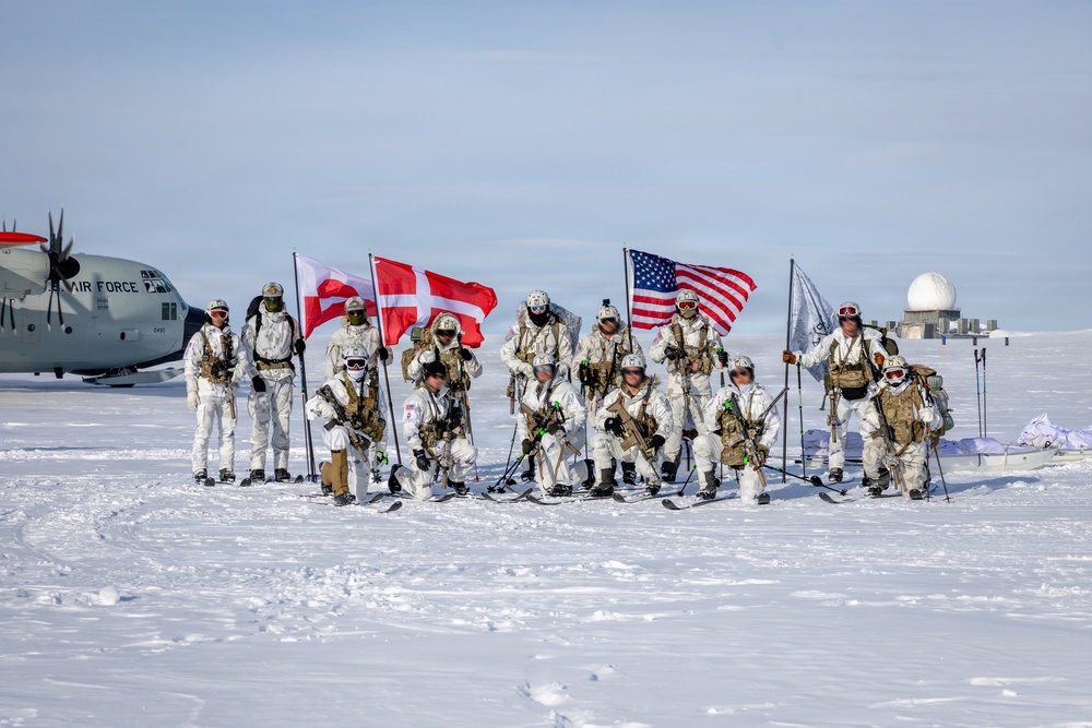 A group of approximately 12-13 military personnel in white arctic/winter camouflage uniforms pose for a group photo on a snow-covered landscape. They are holding multiple flags including the United States flag, the Danish flag, and what appears to be the Greenlandic flag. A military transport aircraft marked 'AIR FORCE' and a radar dome structure are visible in the background.