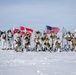 A group of approximately 12-13 military personnel in white arctic/winter camouflage uniforms pose for a group photo on a snow-covered landscape. They are holding multiple flags including the United States flag, the Danish flag, and what appears to be the Greenlandic flag. A military transport aircraft marked 'AIR FORCE' and a radar dome structure are visible in the background.