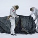 Two military personnel in white winter camouflage uniforms are setting up or dismantling a dark-colored tent in heavy snowfall conditions. Both individuals wear patches on their sleeves that appear to be Danish flags, suggesting they are members of the Danish Armed Forces. The scene depicts cold-weather field operations in an arctic or subarctic environment.
