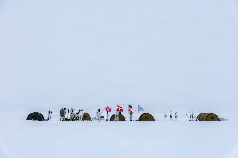 A remote arctic or polar camp is set up on a vast snow-covered landscape under an overcast white sky. Several personnel in white winter camouflage gear are gathered around low-profile tents with skis planted upright in the snow. Multiple national flags — identifiable as Greenland, Denmark, the United States, and at least one other — are displayed on poles, suggesting a multinational military or expedition operation.