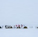 A remote arctic or polar camp is set up on a vast snow-covered landscape under an overcast white sky. Several personnel in white winter camouflage gear are gathered around low-profile tents with skis planted upright in the snow. Multiple national flags — identifiable as Greenland, Denmark, the United States, and at least one other — are displayed on poles, suggesting a multinational military or expedition operation.