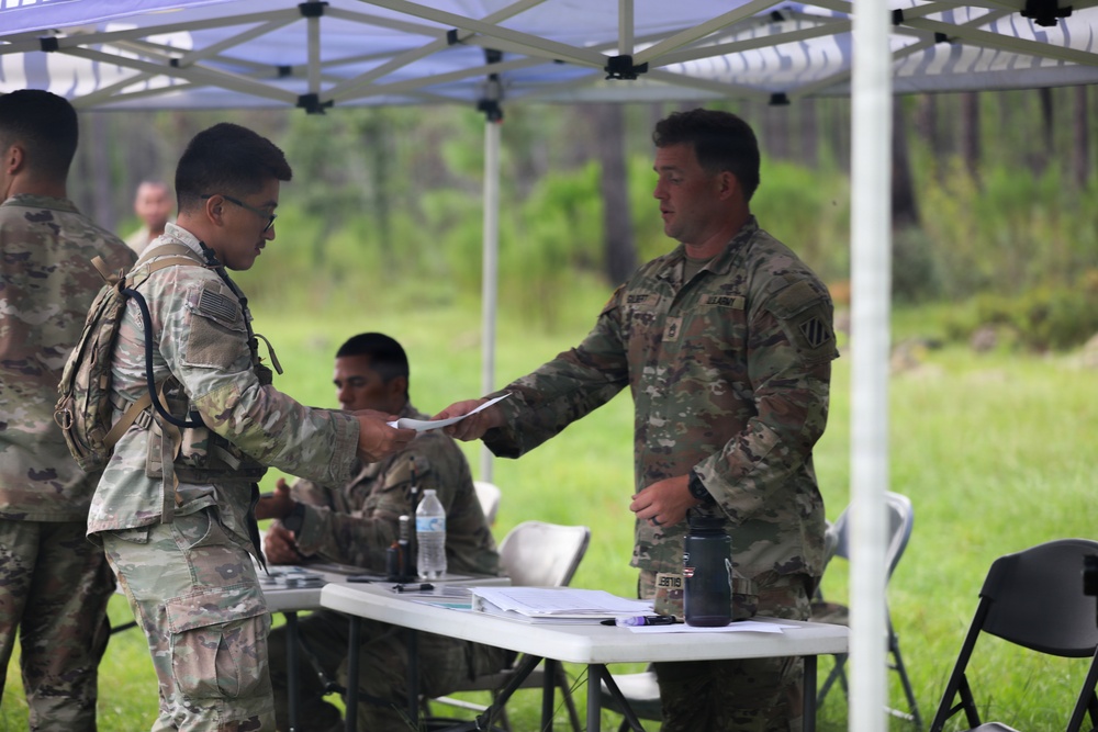 A group of U.S. Army soldiers are gathered under a white pop-up canopy tent in an outdoor field setting. Two soldiers in the foreground are exchanging documents across a folding table, while a third soldier is seated behind the table. The setting appears to be a field administrative checkpoint or processing station during a training exercise.