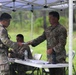 A group of U.S. Army soldiers are gathered under a white pop-up canopy tent in an outdoor field setting. Two soldiers in the foreground are exchanging documents across a folding table, while a third soldier is seated behind the table. The setting appears to be a field administrative checkpoint or processing station during a training exercise.