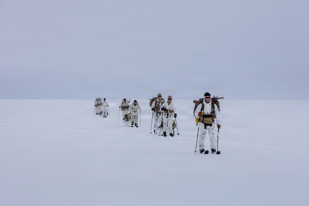A column of military personnel in white winter camouflage uniforms are skiing across a vast, flat, snow-covered terrain under an overcast sky. They are equipped with ski poles, heavy backpacks, and what appear to be rifles slung across their backs. The formation stretches into the distance, suggesting a long-range arctic patrol or cold-weather military exercise.