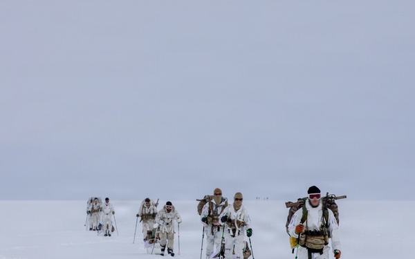 A column of military personnel in white winter camouflage uniforms are skiing across a vast, flat, snow-covered terrain under an overcast sky. They are equipped with ski poles, heavy backpacks, and what appear to be rifles slung across their backs. The formation stretches into the distance, suggesting a long-range arctic patrol or cold-weather military exercise.