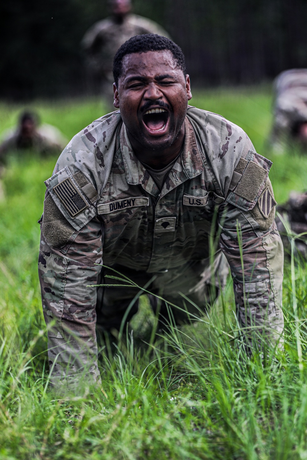 A U.S. Army soldier named Dumeny is captured mid-shout while crawling through tall grass during what appears to be a physical training or field exercise. The soldier is wearing OCP (Operational Camouflage Pattern) uniform with a U.S. flag patch on the left sleeve and is visibly exerting intense effort. Additional soldiers and a wooded background are visible in the out-of-focus background.