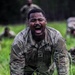 A U.S. Army soldier named Dumeny is captured mid-shout while crawling through tall grass during what appears to be a physical training or field exercise. The soldier is wearing OCP (Operational Camouflage Pattern) uniform with a U.S. flag patch on the left sleeve and is visibly exerting intense effort. Additional soldiers and a wooded background are visible in the out-of-focus background.