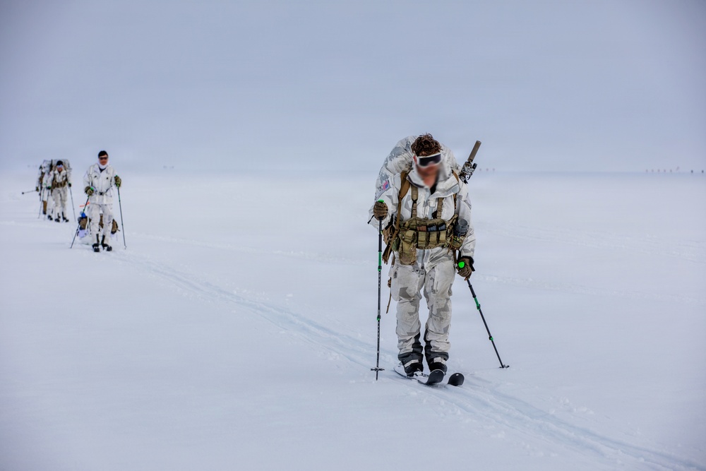 A group of military personnel in white winter camouflage uniforms are cross-country skiing across a flat, snow-covered terrain in low-visibility conditions. The lead figure in the foreground is equipped with tactical gear including a chest rig, large backpack, ski poles, and skis. The group appears to be conducting a cold-weather military training exercise or patrol operation.