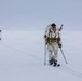 A group of military personnel in white winter camouflage uniforms are cross-country skiing across a flat, snow-covered terrain in low-visibility conditions. The lead figure in the foreground is equipped with tactical gear including a chest rig, large backpack, ski poles, and skis. The group appears to be conducting a cold-weather military training exercise or patrol operation.