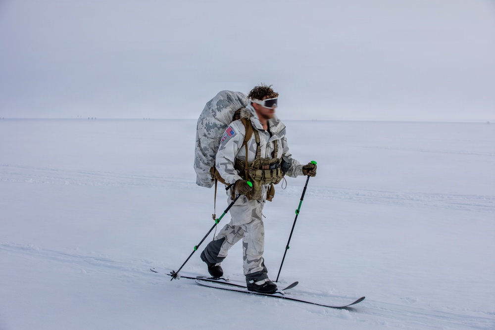 A military personnel in white winter camouflage gear is cross-country skiing across a flat, snow-covered arctic or tundra landscape. The individual is fully equipped with a tactical chest rig, large rucksack, ski poles with green tips, and protective goggles. The overcast, featureless white terrain suggests extreme cold-weather military training or operations.